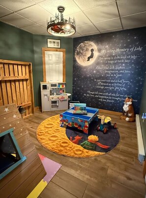 Una habitación acogedora con una zona de juegos de madera, una estantería, un microondas y un mural en memoria de alguien.