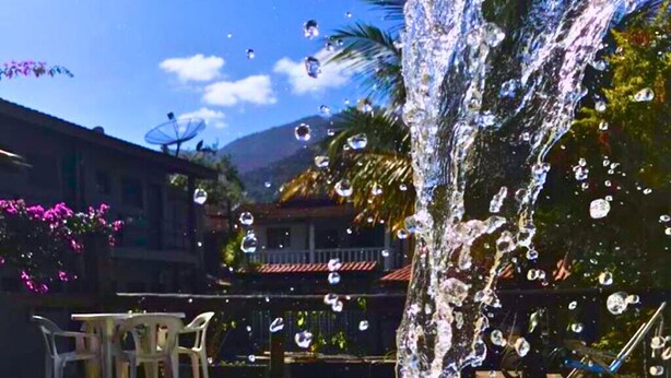 A pool with water spouting from a fountain, surrounded by chairs and a building with a tiled roof. - VEREDA 7 SUITES (São Sebastião)