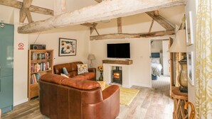 A rustic living room with exposed wooden beams, a leather sofa, a bookshelf, a fireplace, and a television. - Megs Cottage (Stratford-upon-Avon)