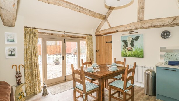 A dining area with a wooden table and chairs, a clock on the wall, and a view through a glass door. - Megs Cottage (Stratford-upon-Avon)