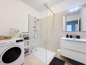 Ein modernes Badezimmer mit Duschkabine aus Glas, einer Waschmaschine, einem Waschtisch mit Spiegel und einem kleinen Ablagefach für Pflegeprodukte.