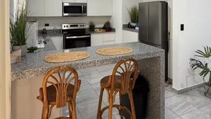 A modern kitchen with white cabinets, a stainless steel microwave, a built-in oven, a granite countertop, two wooden bar stools, and a refrigerator.