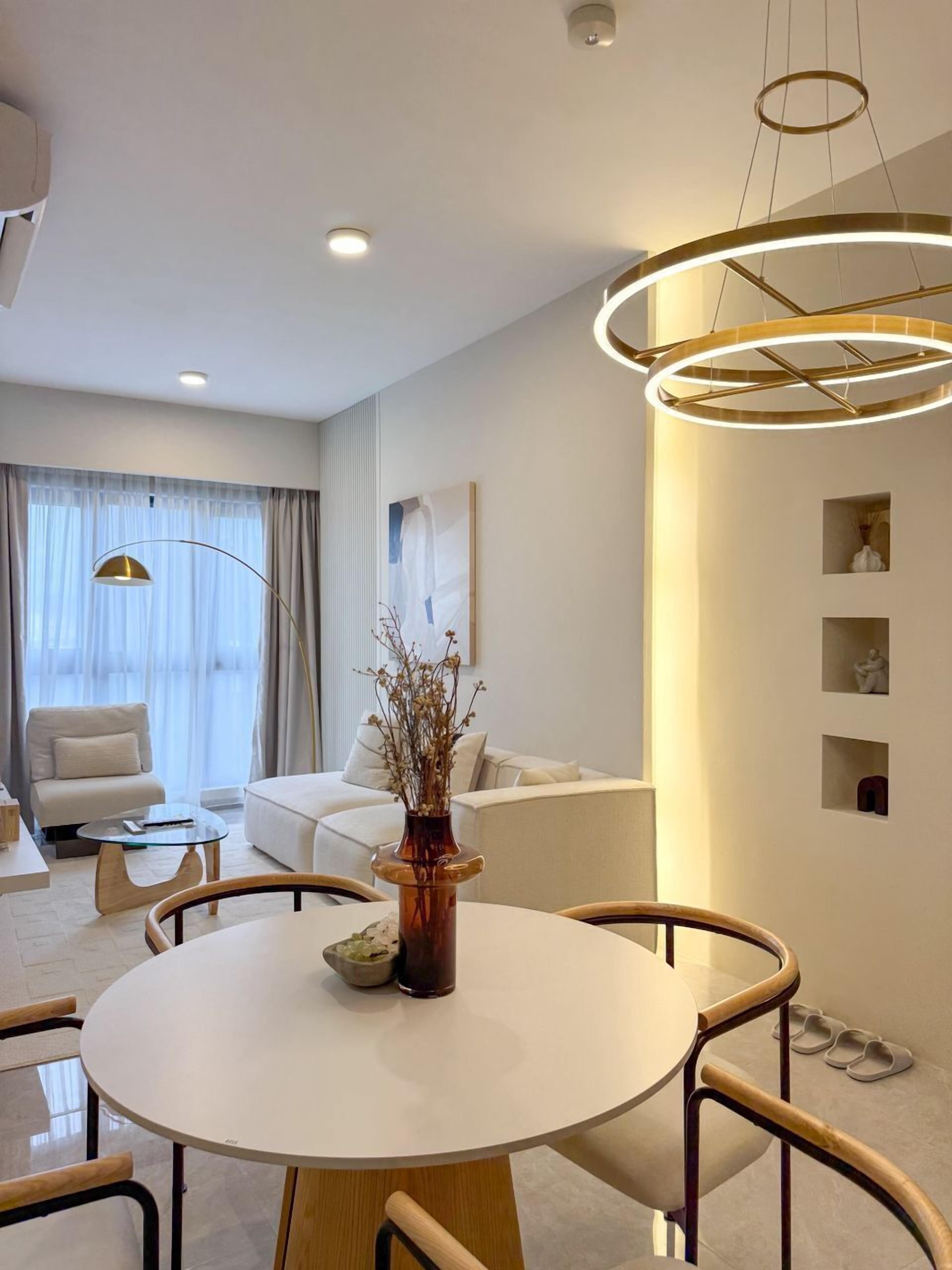 Quarto de hotel moderno com uma mesa de jantar redonda, um sofá, uma cadeira, um vaso com flores secas e uma luminária pendente.