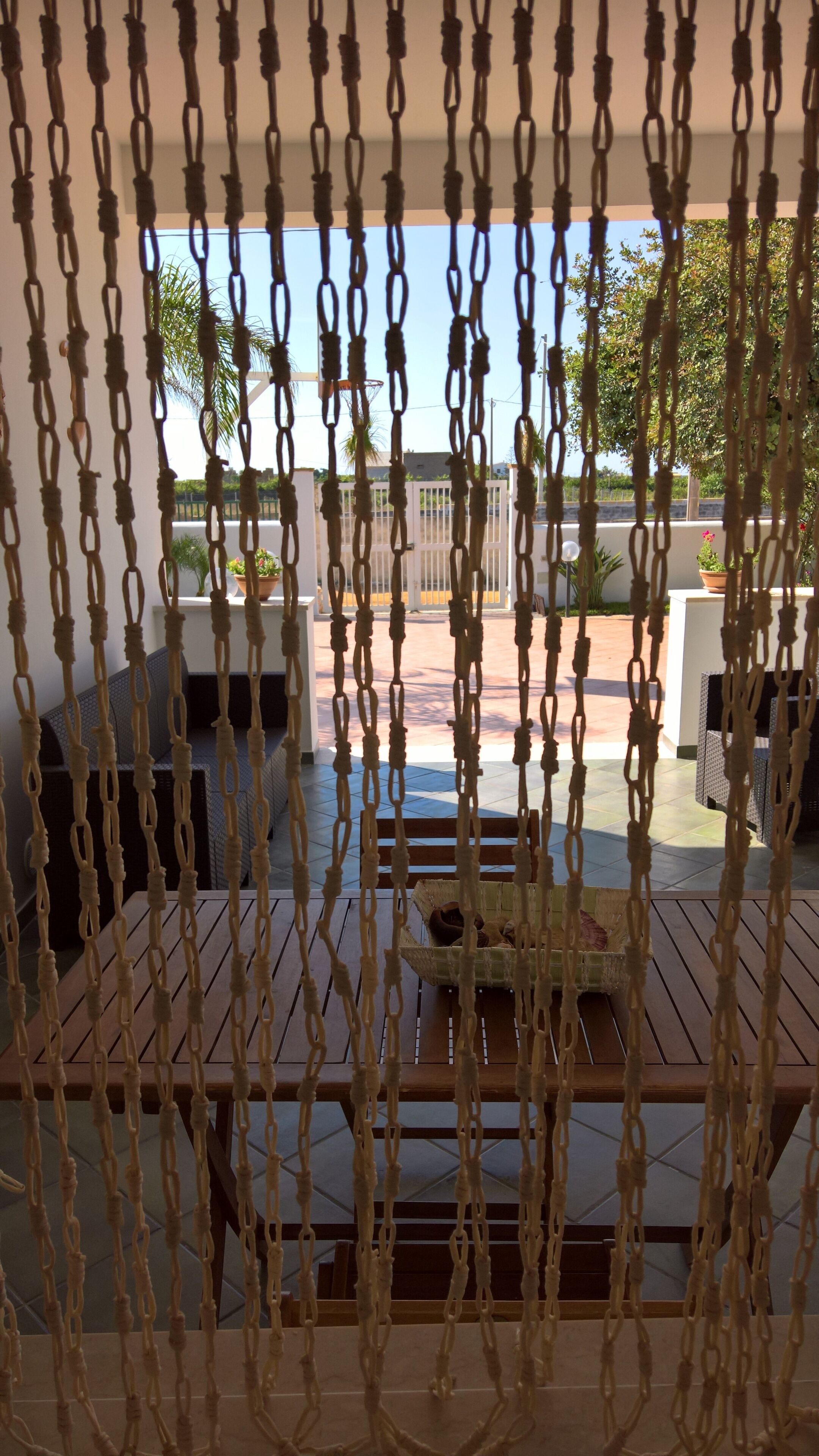 A view through macrame hanging decor to an outdoor patio area with a wooden table and chairs, a white fence, and greenery in the background.
