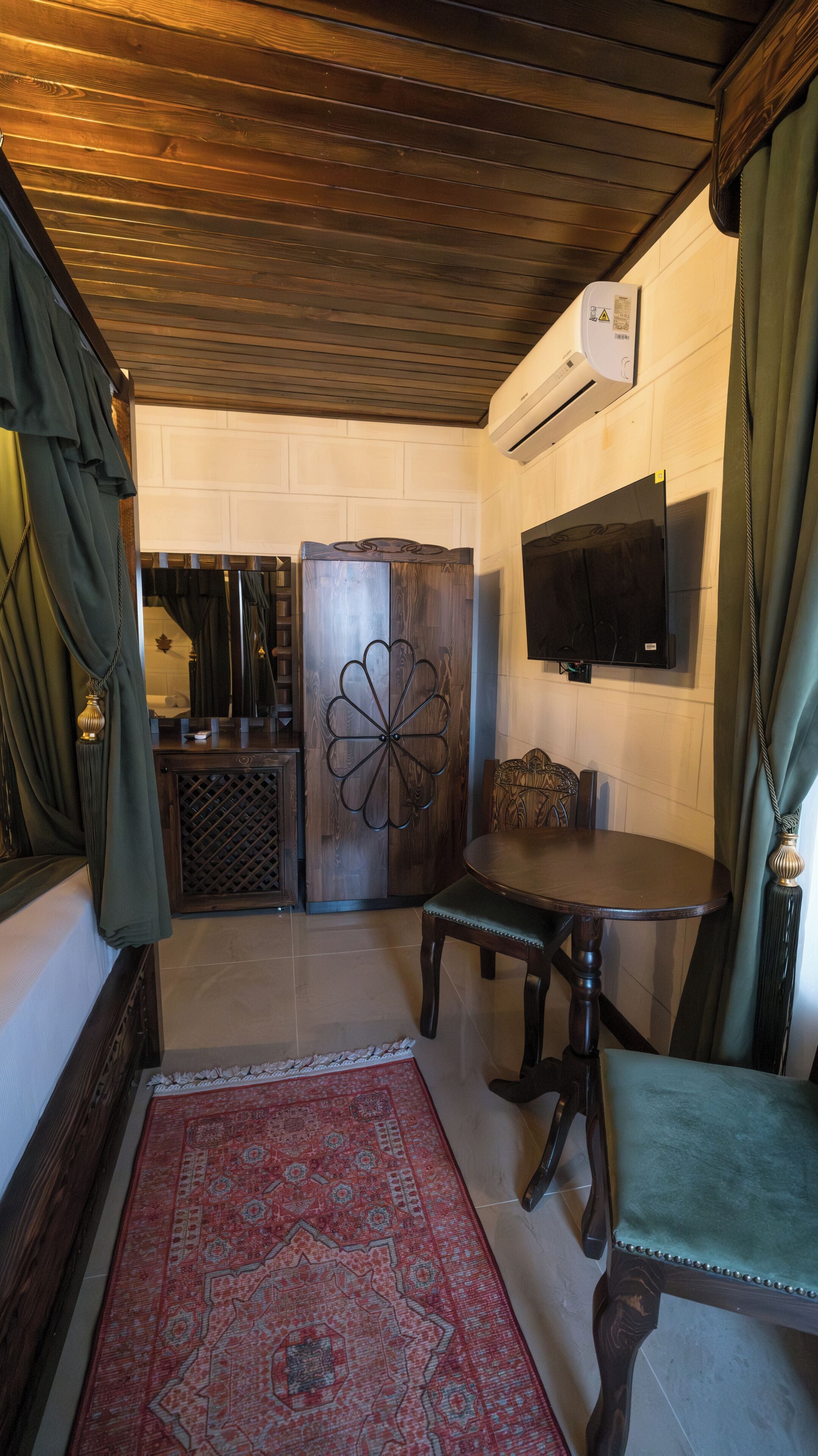 A room with a wooden ceiling, a TV mounted on the wall, a dining table with chairs, and a patterned rug.