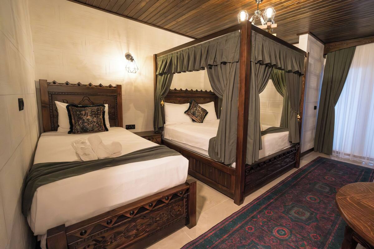 A four-poster bed with a canopy, a separate bed with a headboard, a wooden table, and a patterned rug.