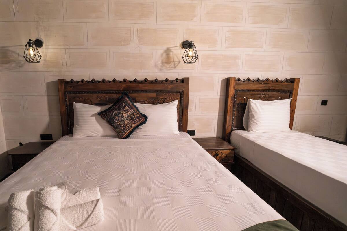 A hotel room with a large bed, two bedside tables, a wooden headboard, a decorative pillow, and wall-mounted lights.