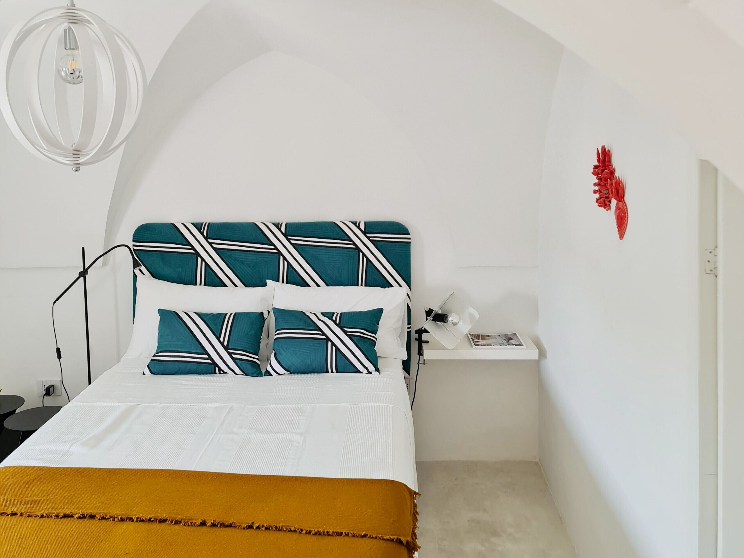 A bedroom with a white bed, teal and white patterned headboard, a mustard-colored throw, a white nightstand with a lamp, and a red wall decoration.