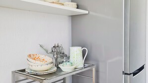 Una cocina con un refrigerador de acero inoxidable, una repisa de vidrio con vajilla y una mesa de metal con cuencos y utensilios.