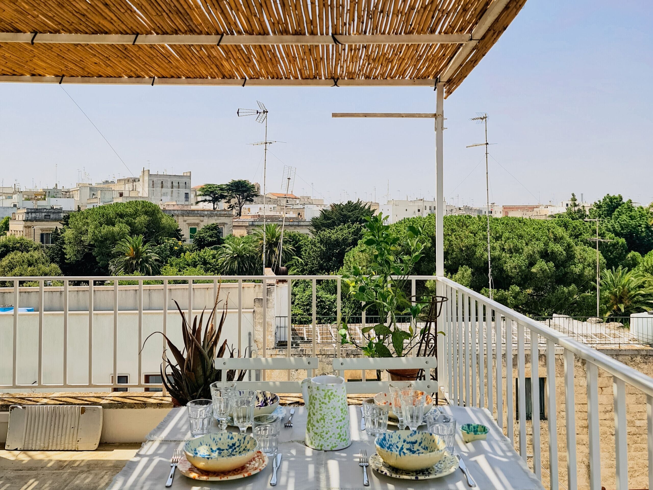 An outdoor dining area with a table set for two, surrounded by greenery and overlooking a cityscape.