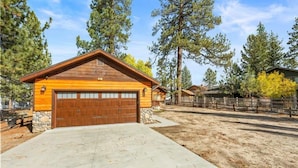 Une cabane en bois avec un garage, entourée d’arbres et d’une zone délimitée par une clôture.