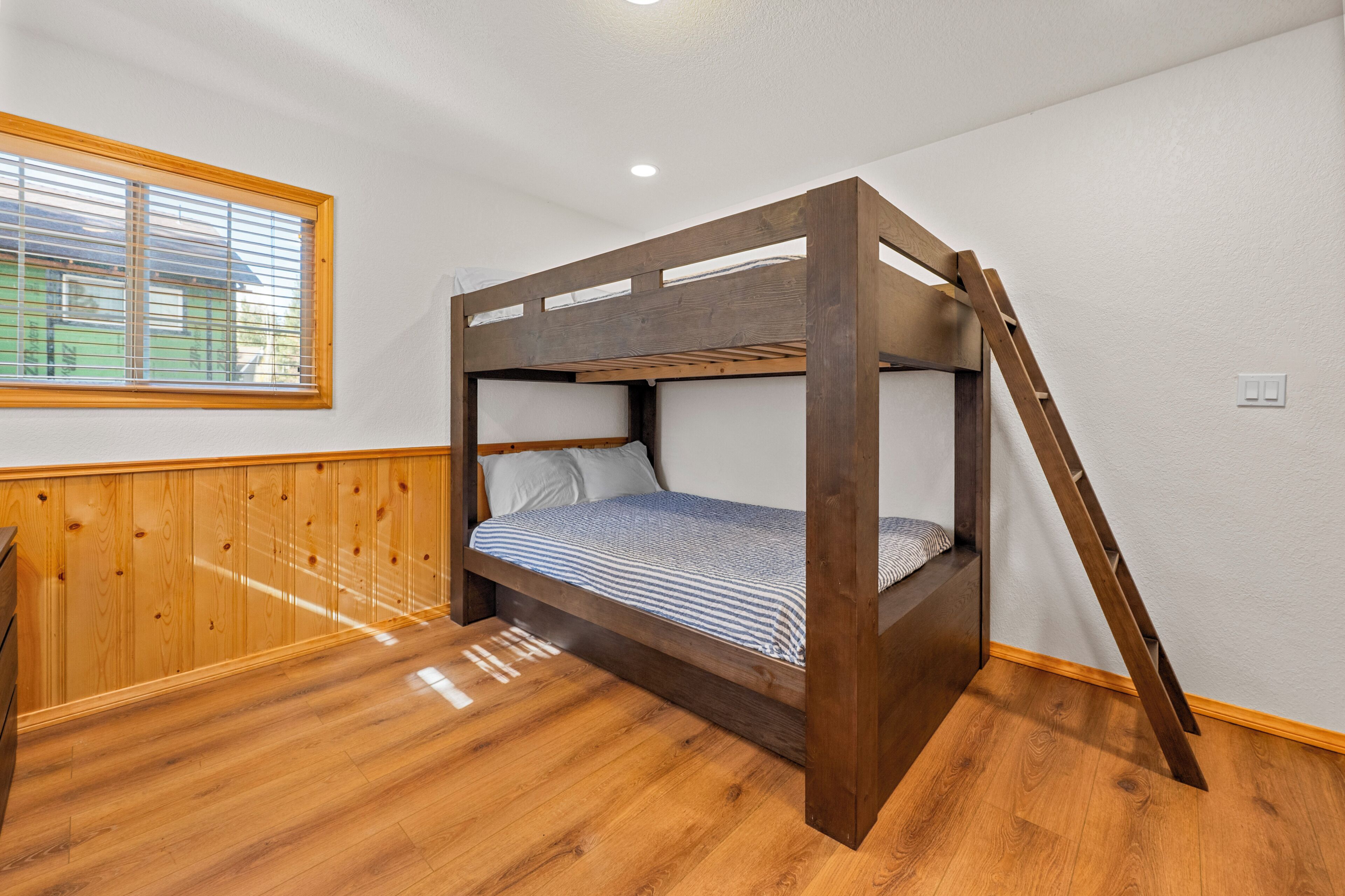A bunk bed with a wooden ladder, a window with blinds, and a wooden floor.