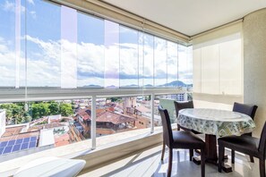 A balcony with a table and chairs, a view of a cityscape, and a large window. - Apartamento Atalaya 83 (Guarujá)