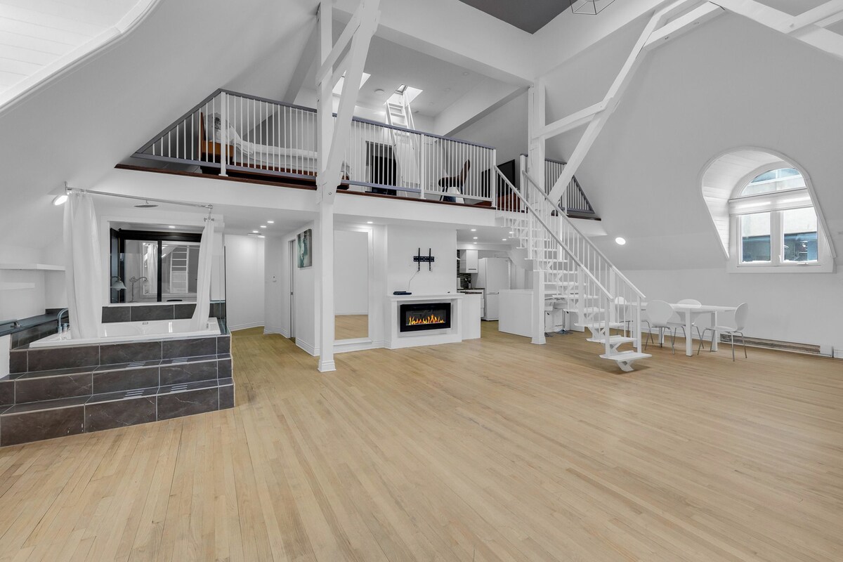 A spacious loft with a high ceiling, a fireplace, a staircase, and a balcony.