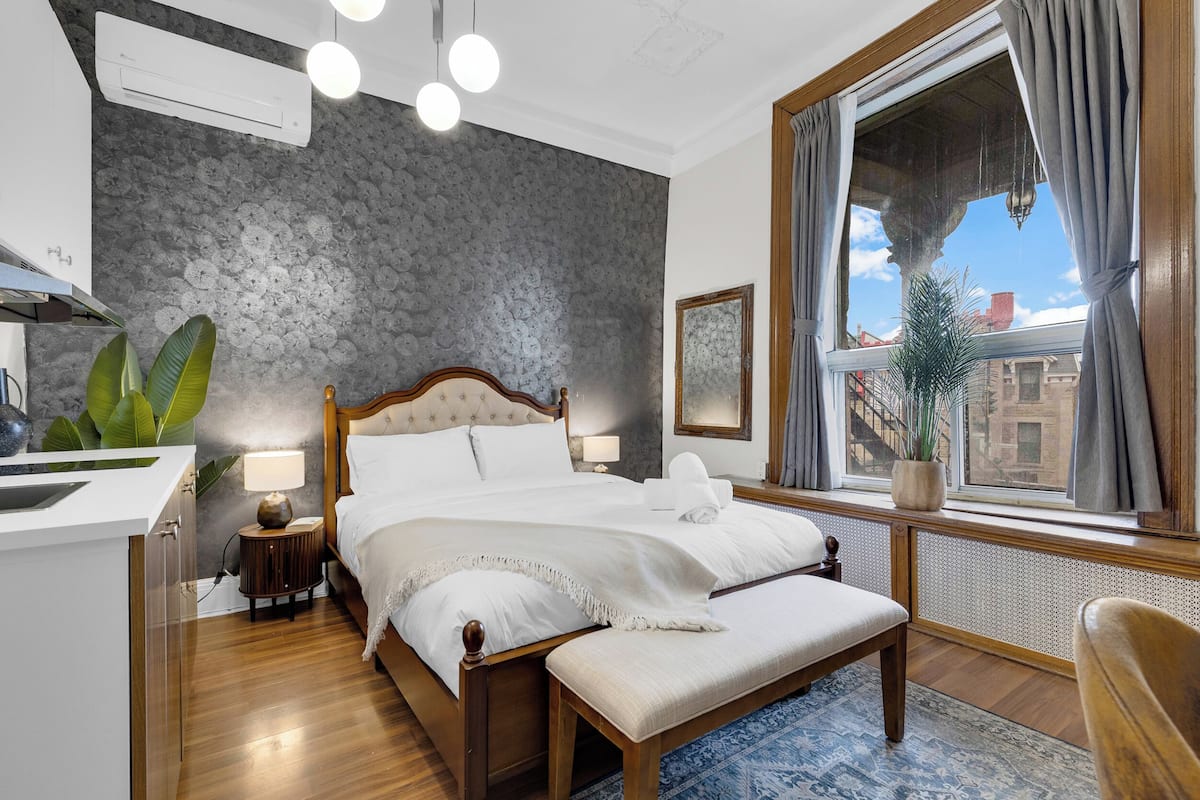 Un dormitorio con cama, mesita de noche, lámpara, planta en maceta, ventana con cortinas y vistas a edificios.