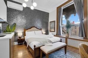 A bedroom with a bed, bedside table, lamp, potted plant, window with curtains, and a view of buildings. - Maison Paris (Montréal)