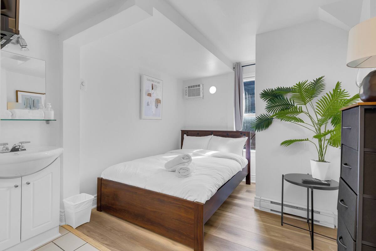 Un dormitorio con una cama de madera, ropa de cama blanca, una mesita de noche con una planta, un armario blanco y un ventanal grande.