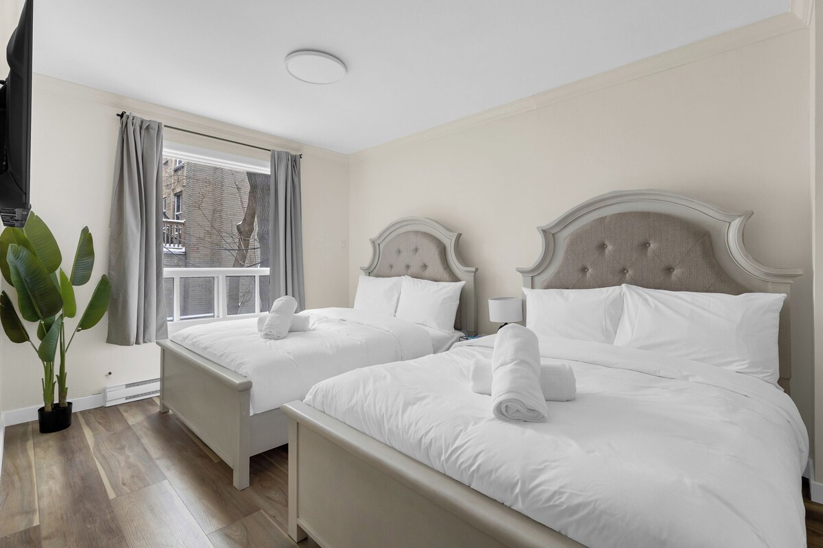 A hotel room with two beds, a window with curtains, a potted plant, and a ceiling light.