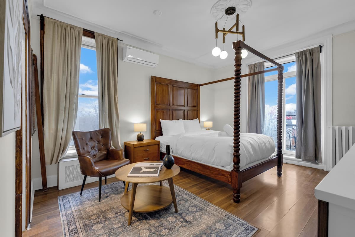 Una cama con dosel de madera, un sillón de cuero, una mesita de noche con lámpara, y vistas a la ciudad a través de amplios ventanales.