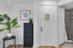 A white door with a security panel, a framed abstract painting, a potted plant, a black filing cabinet, and a small table with a lamp. - Maison Paris (Montréal)