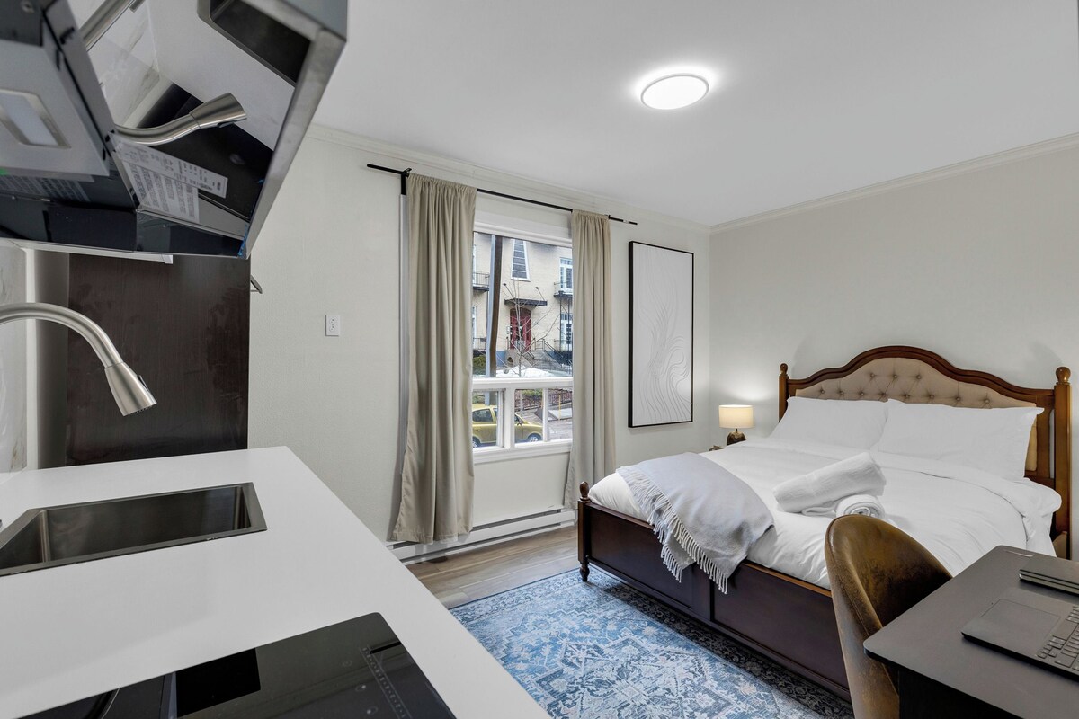 A hotel room with a bed, a desk with a laptop, a window with curtains, and a view of buildings outside.