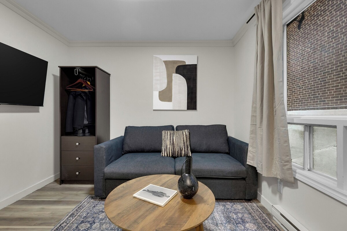 A modern living room with a gray sofa, a round coffee table, a black vase, a hanging artwork, a wardrobe, and a window with curtains.