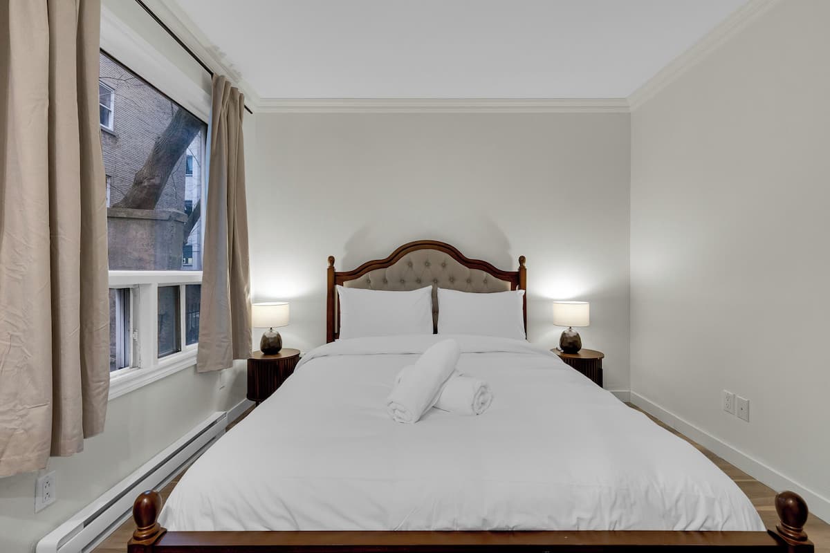 Una cama bien hecha con sábanas blancas, dos lámparas de noche y una ventana con cortinas.