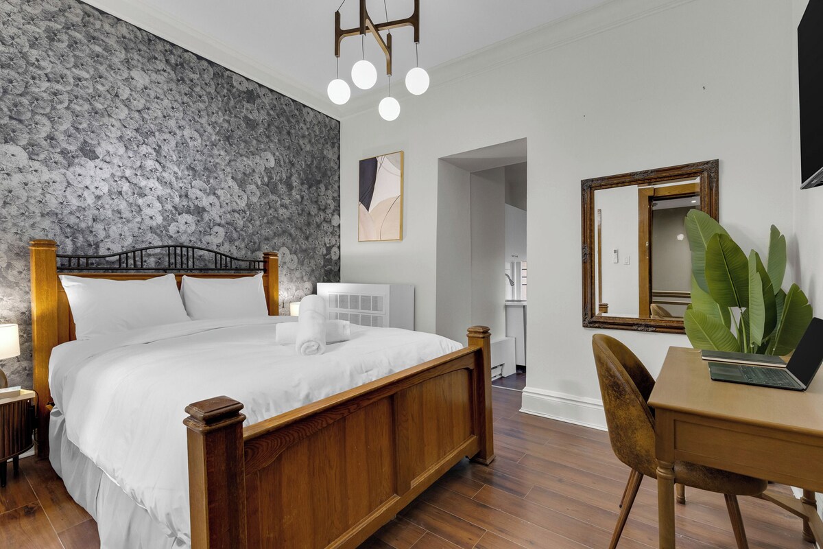 A hotel room with a wooden bed, a desk with a laptop, a large mirror, and a plant.