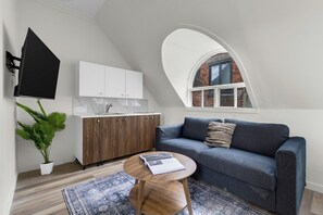 A modern loft apartment with a kitchen, a living area with a sofa, a round wooden table, and a potted plant. - Maison Paris (Montréal)