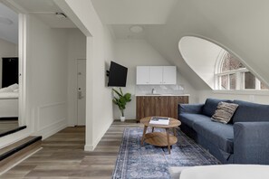 A modern loft bedroom with a large window, a flat-screen TV, a wooden cabinet, and a blue patterned rug. - Maison Paris (Montréal)