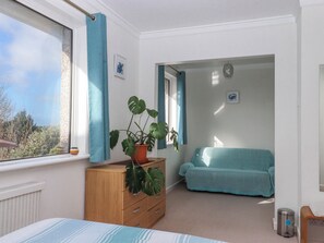 Ein Schlafzimmer mit einem Bett, einer Holzkommode, einem Topfpflanzen, einem salbeigreenen Sofa und einem Fenster mit Vorhängen.