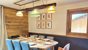 Un coin repas avec une table en bois dressée pour six personnes, des chaises bleues et une chaise rose. On y trouve trois lampes suspendues, des poutres en bois au plafond et des tableaux encadrés au mur.