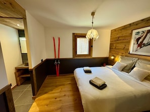 Un dormitorio con suelo de madera, una cama con sábanas blancas, una ventana con persianas, una lámpara colgante y un cabecero de madera.