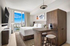 A hotel room with a bed, a desk with two stools, a wardrobe, a television, and a large window offering a city view. - Domus Brickell Studio Suite (Miami)