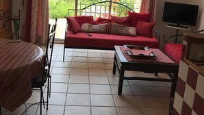 Ein Wohnzimmer mit einer Couch, einem Tisch, einem Fernseher und Blick auf einen Garten durch große Fenster.