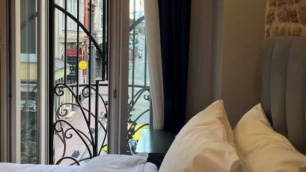 Habitación de hotel con cama, vista de la calle a través de una ventana con baranda metálica y una lámpara fijada a la pared.