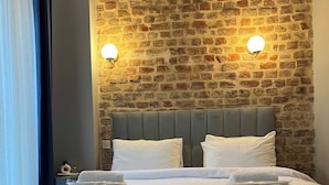 Una cama con cabecera gris, almohadas blancas y toallas dobladas. Se aprecia una pared de piedra con diseño de ladrillo y dos luces fijadas a la pared.