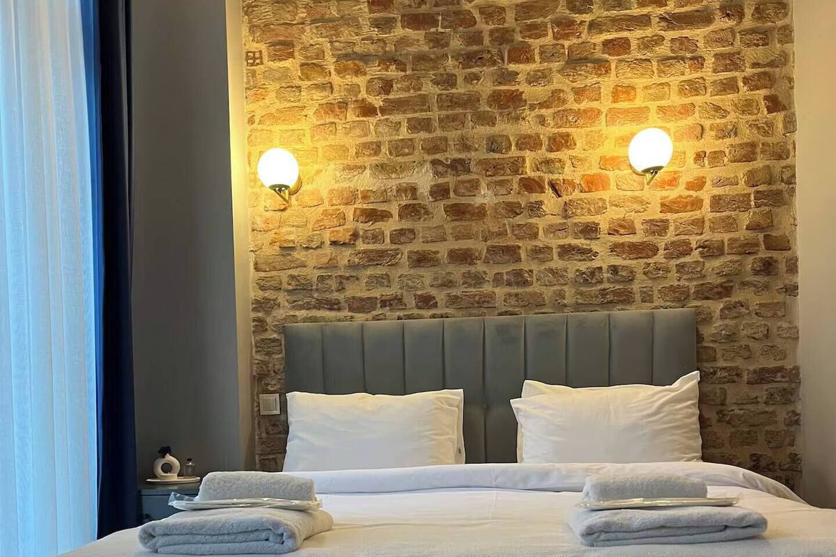 A bed with a grey headboard, white pillows, and folded towels. There is a stone wall with a brick pattern and two wall-mounted lights.