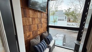 A room with a wooden wall, a sofa with striped cushions, a mounted TV, and a view of a house through a large window. - Harbor Lights Resort Waterfront Pool Dock (Oak Island)
