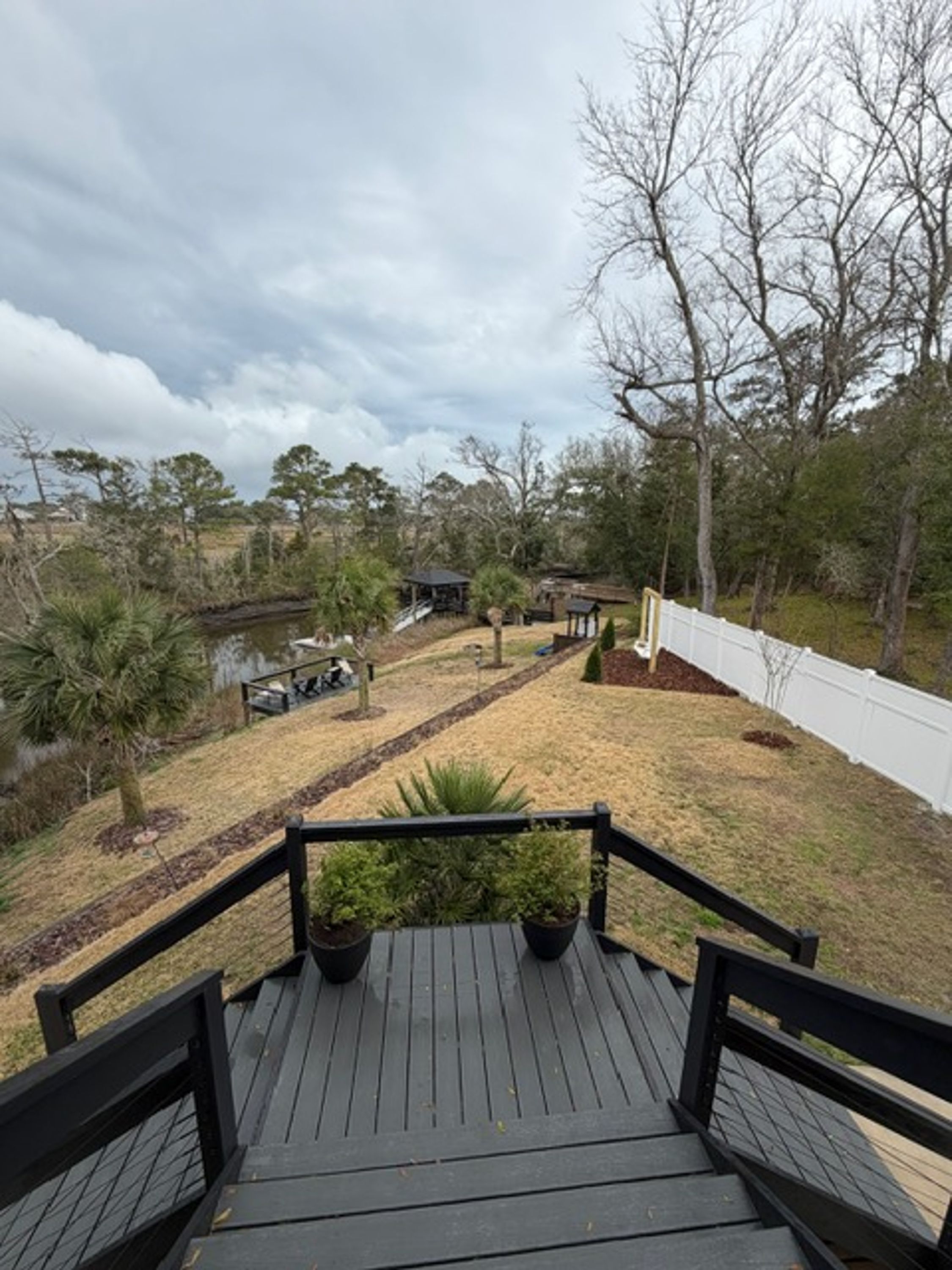 A wooden deck with a view of a backyard, including a pond, trees, and a white fence.