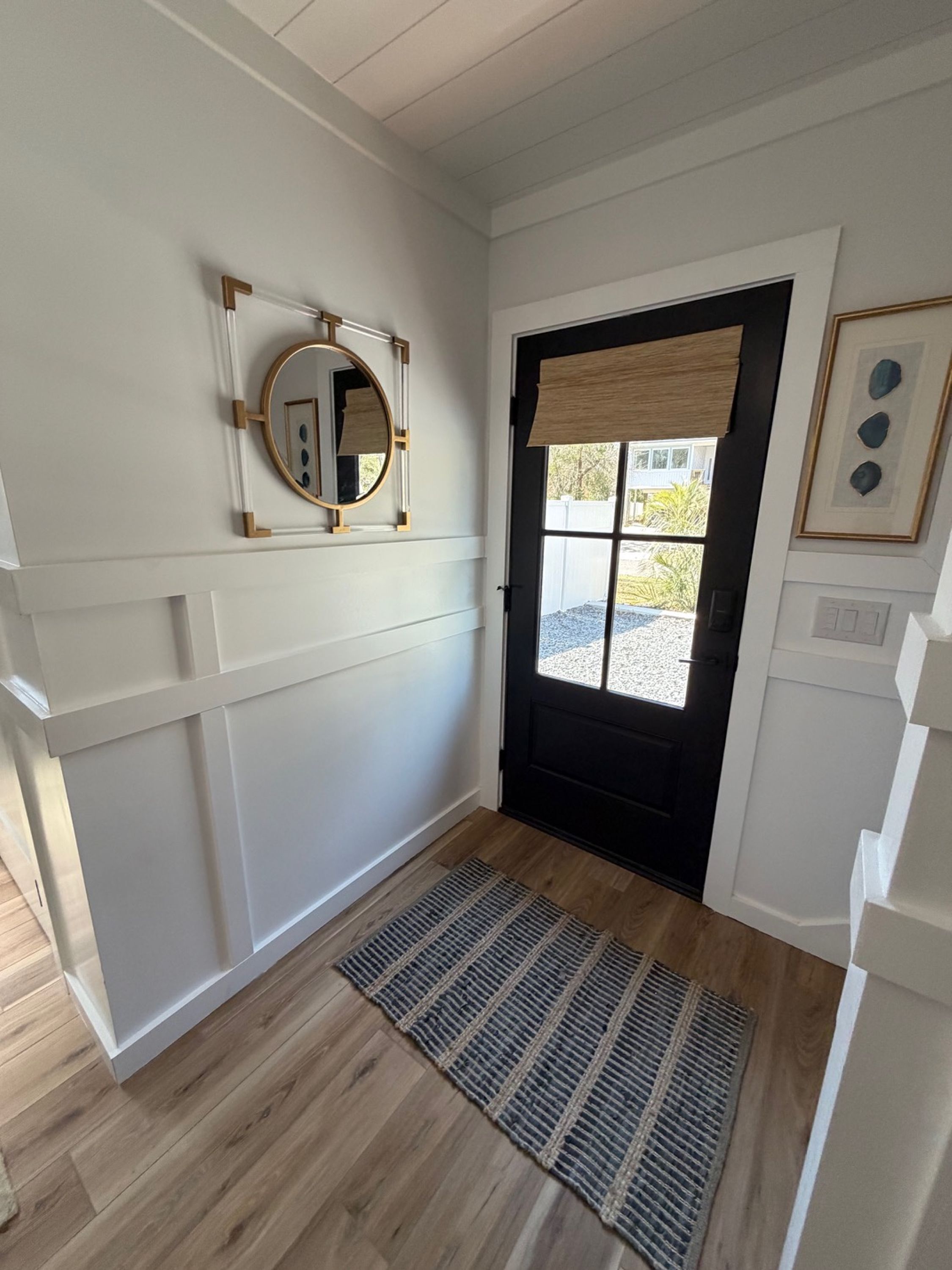 A hallway with a black door, a white wall with a mirror, and a wooden floor.