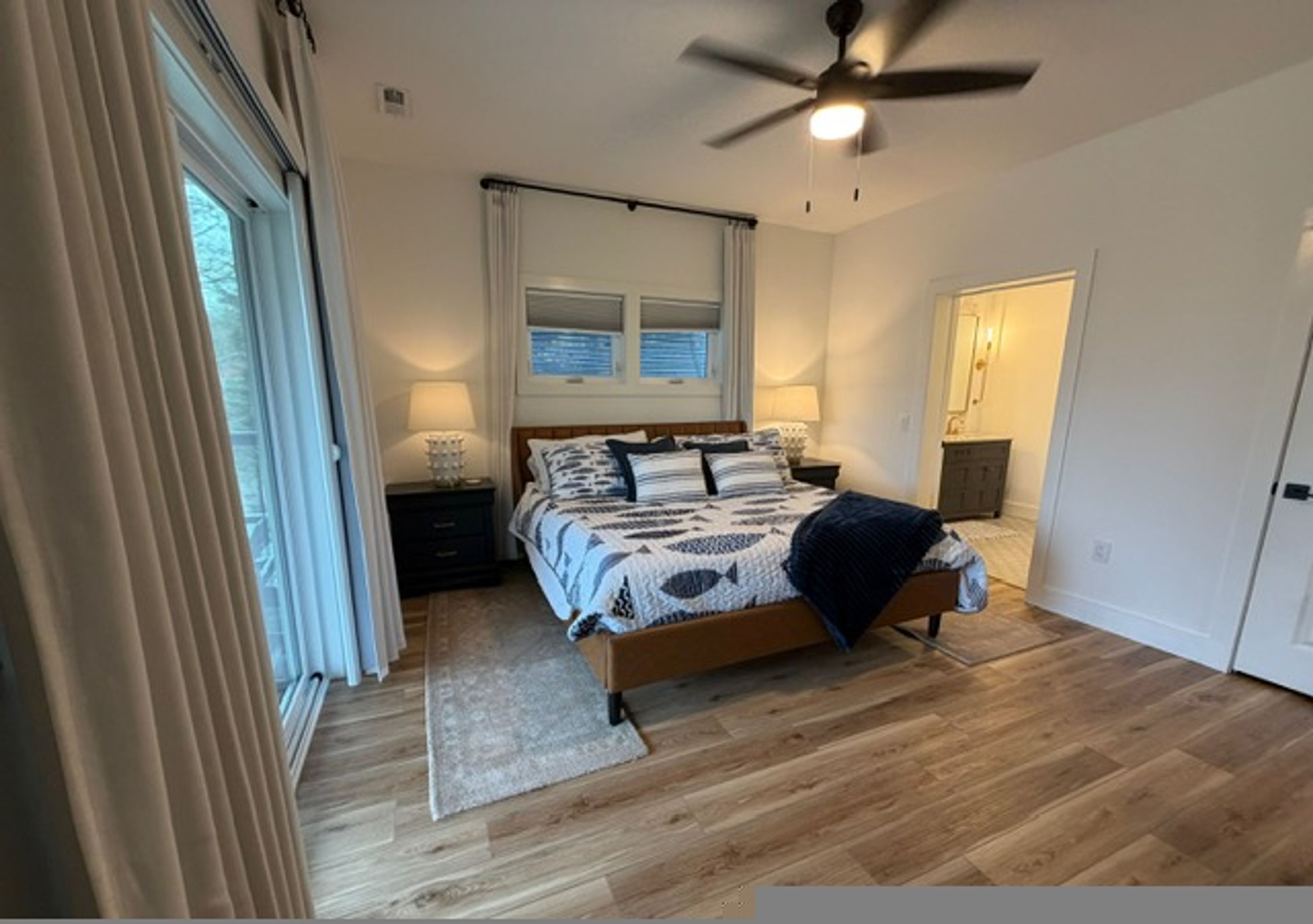 A bedroom with a bed, a ceiling fan, a window with curtains, a nightstand, and a door.