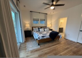 Un dormitorio con cama, ventilador de techo, ventana con cortinas, mesita de noche y una puerta.