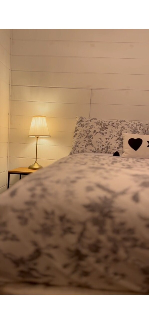 A bed with a floral bedspread, a nightstand with a lamp, and a wall with a floral-patterned pillow.