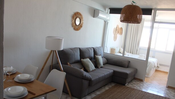 Ein modernes Wohnzimmer mit einer grauen Couch, weißen Stühlen, einem Esstisch mit Tellern, einer Stehlampe und einem großen Fenster mit Vorhängen.