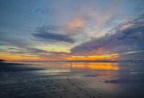 A beach with footprints leading to the water, a sunset in the background, and a calm sea. - Ocean-View Spa Home / Sauna · Cold Plunge · Hot Tub / Sleeps 10 (Long Beach)