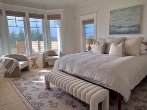 A bedroom with a large bed, a bench, a chair, and a view of the outdoors through large windows. - Ocean-View Spa Home / Sauna · Cold Plunge · Hot Tub / Sleeps 10 (Long Beach)