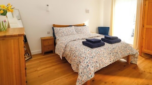 A bedroom with a wooden bed, a nightstand, a vase with flowers, a chair, and a wooden wardrobe. - The Pebble (Dorchester)
