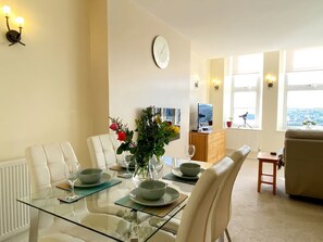 A dining area with a glass table, white chairs, a clock, and a view of the city through large windows. - Sea View Apartment, 7 Ellington Court (Torquay)