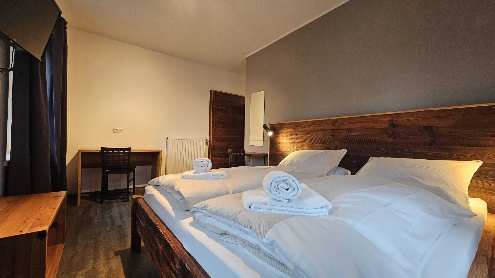 A double bed with white linens and rolled towels, a wooden headboard, a desk with a chair, and a radiator.